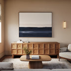 A modern living room features a geometric wooden sideboard, a low wooden coffee table, a light gray sofa, a textured rug, and the Ivory Dusk Canvas Art – Horizontal in neutral and dark blue tones for a sophisticated touch.