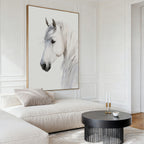 A modern living room with a large white sofa, a round black coffee table, neutral tones, minimal decor, and the Ivory Mane Canvas Art—a framed painting of a white horse with a soft background—on the wall.