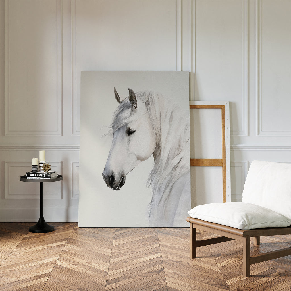 A minimalist room with white paneled walls, wood herringbone floor, a cushioned chair, a small round table with candles and books, and the Ivory Mane Canvas Art—striking white horse print—propped against the wall.
