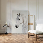 A minimalist room with white paneled walls, wood herringbone floor, a cushioned chair, a small round table with candles and books, and the Ivory Mane Canvas Art—striking white horse print—propped against the wall.