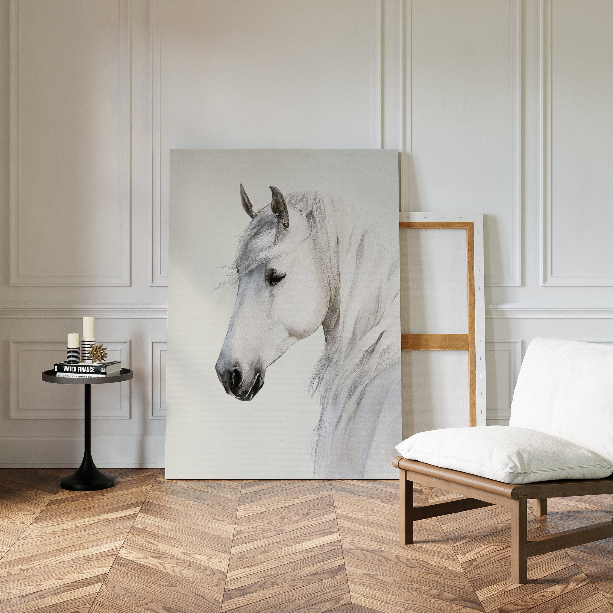 A minimalist room with white paneled walls, wood herringbone floor, a cushioned chair, a small round table with candles and books, and the Ivory Mane Canvas Art—striking white horse print—propped against the wall.