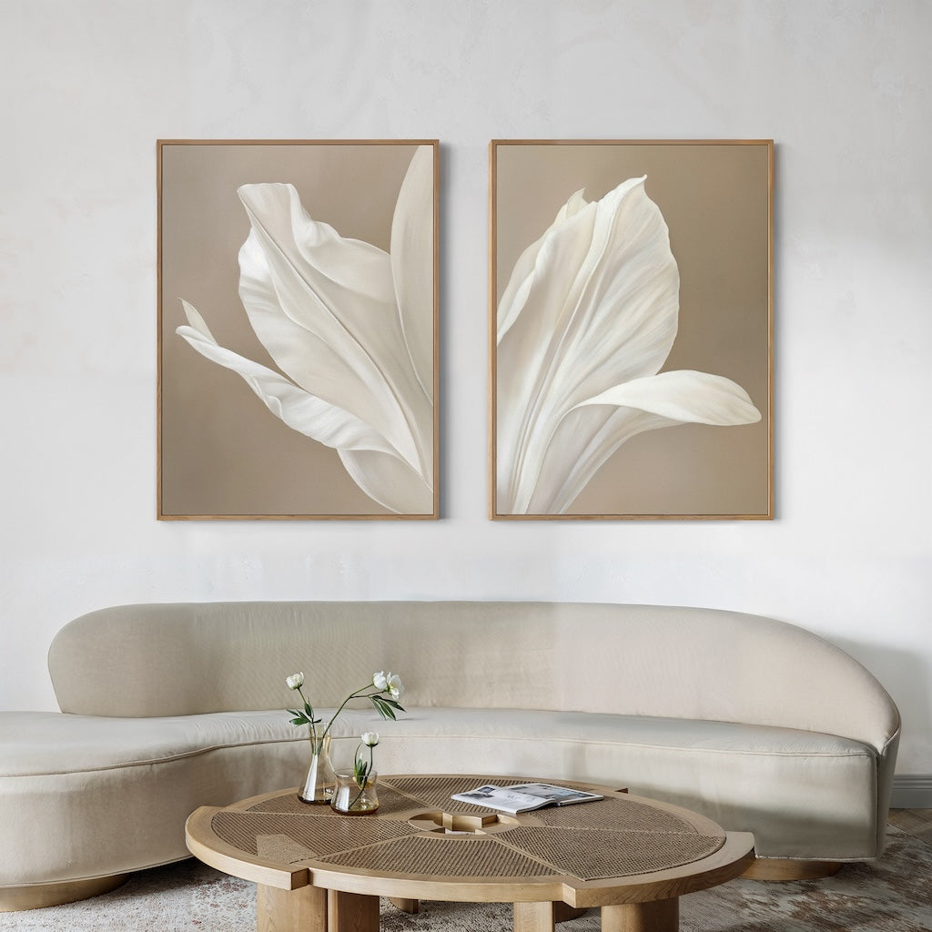 A modern living room features a curved cream sofa, a round wooden coffee table with a glass vase and magazines, and the Ivory Petals Set of 2 wall art—showcasing white petals on beige walls for stylish minimalist decor.