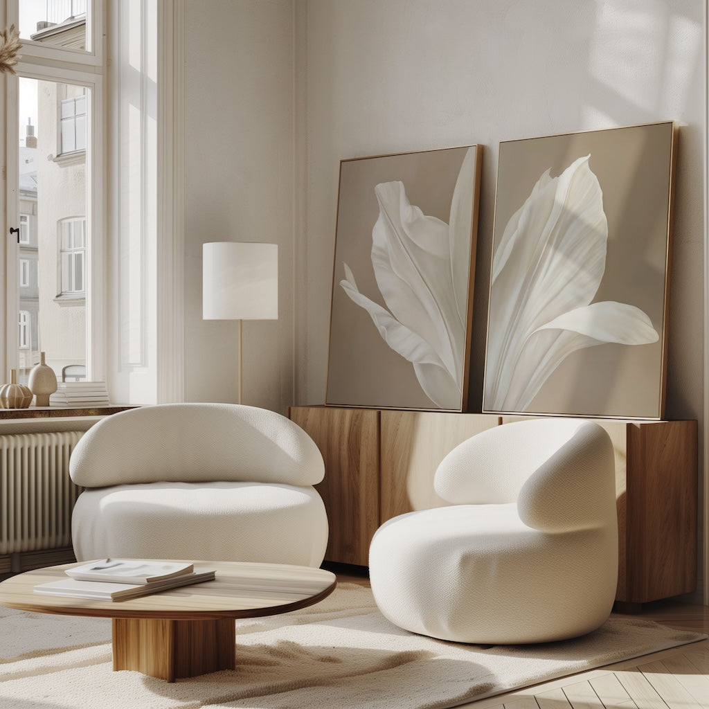 A modern living room with two curved white armchairs, a wooden coffee table, a floor lamp, and the Ivory Petals Set of 2 wall art on a sideboard under tall windows—perfect for chic decor filled with natural light.