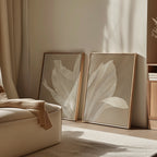 The Ivory Petals Set of 2, featuring beige and white tones, leans against the wall in a softly lit, minimalist room. A cream chair with a tan throw completes the cozy and inviting living room decor.