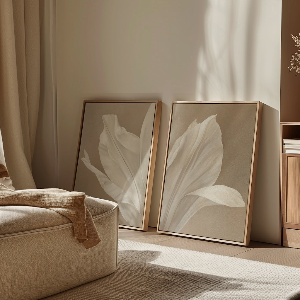 The Ivory Petals Set of 2, featuring beige and white tones, leans against the wall in a softly lit, minimalist room. A cream chair with a tan throw completes the cozy and inviting living room decor.