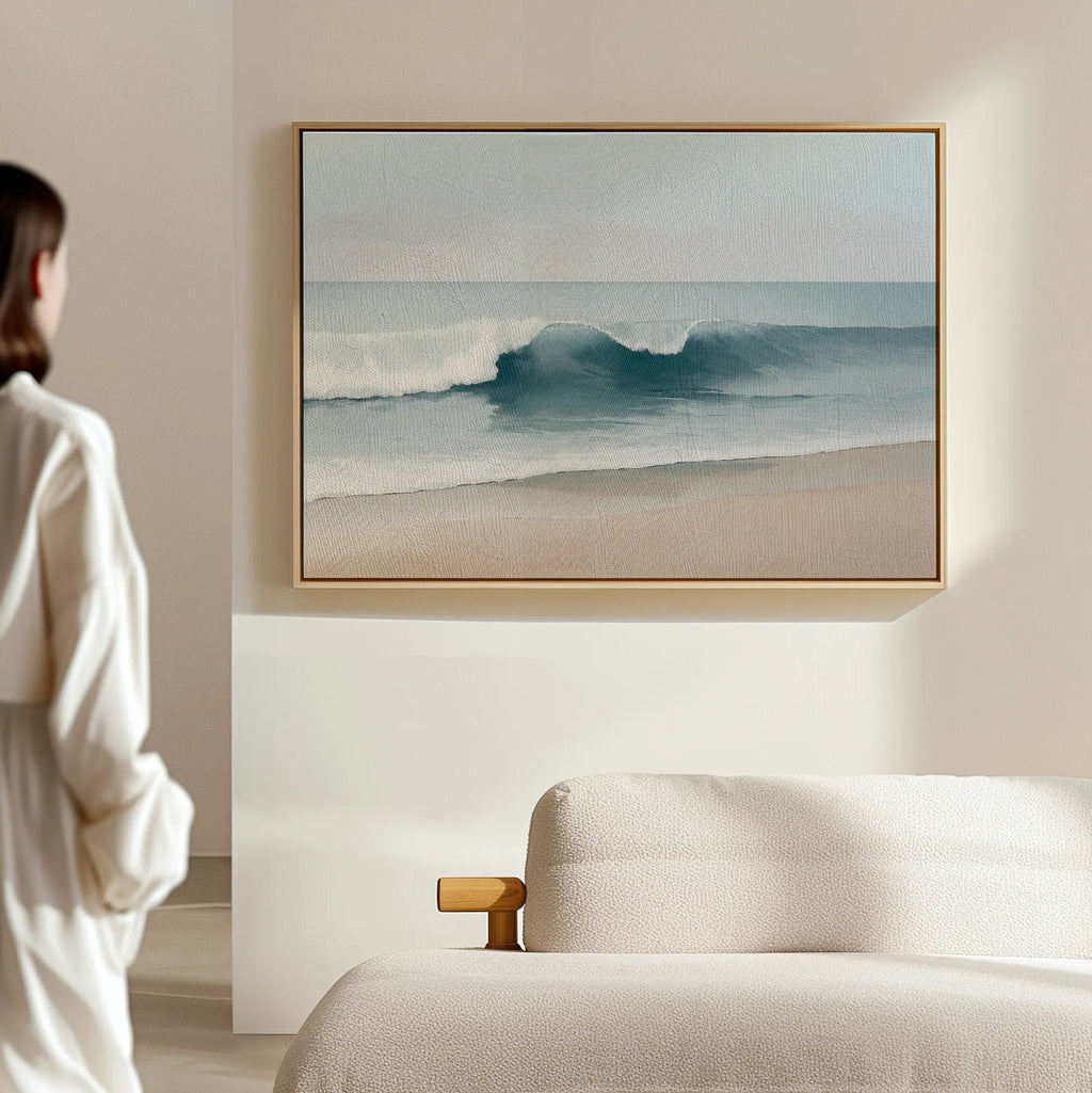 A person in a white shirt stands in a bright, minimal room, gazing at the Lapping Tide Canvas Art—ocean waves wall art—displayed above a beige sofa.