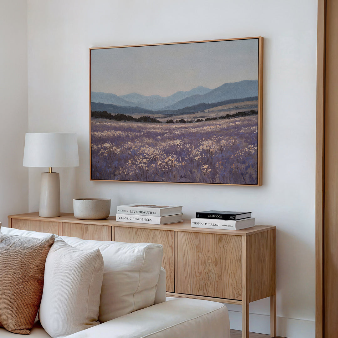 A cozy living room corner with a wooden sideboard, books, lamp, and bowl features Lavender Terrain Canvas Art, a framed landscape of lavender fields and mountains that brings a calming touch to the white wall.