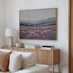 A cozy living room corner with a wooden sideboard, books, lamp, and bowl features Lavender Terrain Canvas Art, a framed landscape of lavender fields and mountains that brings a calming touch to the white wall.