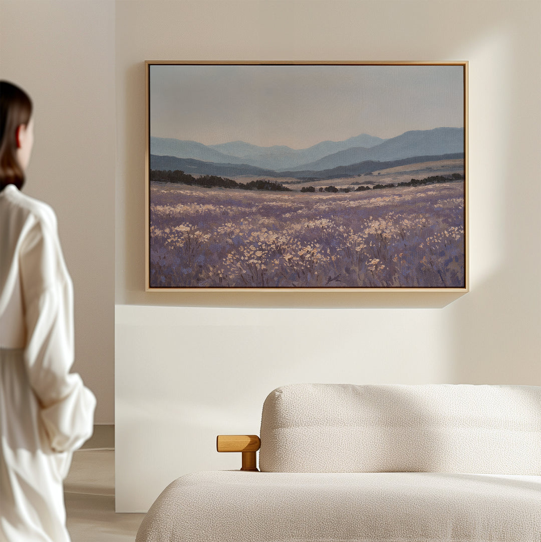 A woman in white admires the Lavender Terrain Canvas Art—a calming print of purple wildflowers and distant mountains—hanging above a beige sofa on a light-colored wall.