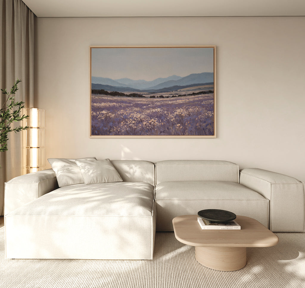 A modern living room with a light beige sectional sofa, wooden coffee table, potted plant, and the Lavender Terrain Canvas Art featuring lavender fields and mountains hanging on a neutral wall. Natural light fills the calming space.