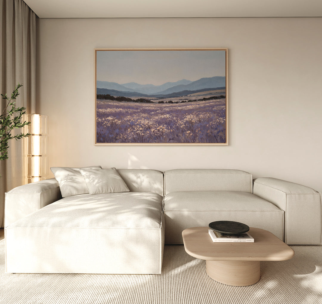 A modern living room with a light beige sectional sofa, wooden coffee table, potted plant, and the Lavender Terrain Canvas Art featuring lavender fields and mountains hanging on a neutral wall. Natural light fills the calming space.