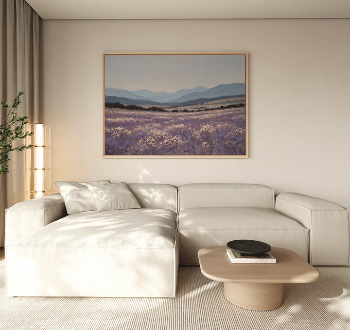 A modern living room with a light beige sectional sofa, wooden coffee table, potted plant, and the Lavender Terrain Canvas Art featuring lavender fields and mountains hanging on a neutral wall. Natural light fills the calming space.
