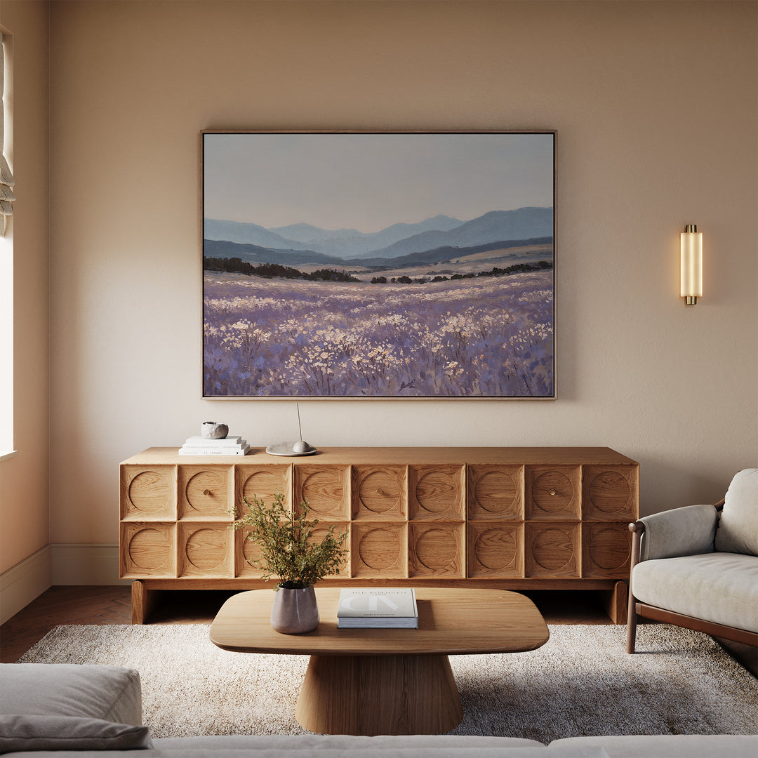 A cozy living room with a light sofa, wooden coffee table, and geometric sideboard. Above the sideboard hangs the Lavender Terrain Canvas Art, a calming wall print. A vase with flowers rests on the coffee table.