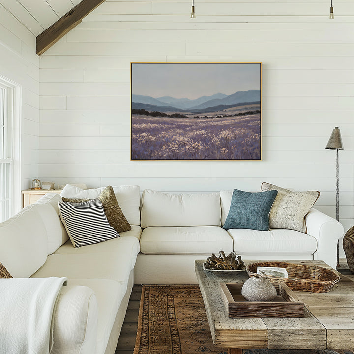A bright, airy living room with white shiplap walls, a white sectional sofa and assorted throw pillows, a wooden coffee table, and the calming Lavender Terrain Canvas Art framed above the sofa.