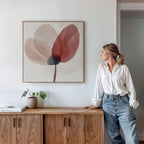 A woman in a white blouse and jeans leans on a wooden cabinet, admiring the Layered Bloom Canvas Art. A small potted plant and book rest atop the cabinet, enhancing the modern wall decor of this serene space.