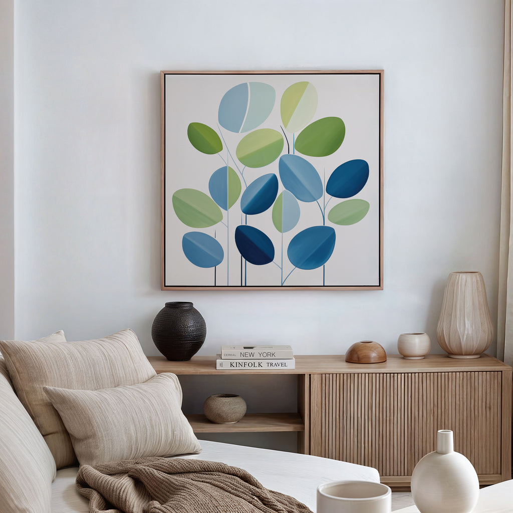 A modern living room features a beige sofa, cushions, a wooden sideboard with vases and books, and minimalist wall art—Layered Stillness Canvas Art—showcasing green and blue leaves. Natural light filters through beige curtains.