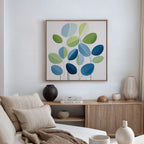 A modern living room features a beige sofa, cushions, a wooden sideboard with vases and books, and minimalist wall art—Layered Stillness Canvas Art—showcasing green and blue leaves. Natural light filters through beige curtains.