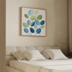 A modern bedroom features a beige upholstered bed with white bedding and four pillows. Above the bed hangs the Layered Stillness Canvas Art, showcasing green and blue leaf shapes. A bare branch sits in a vase on the bedside table.