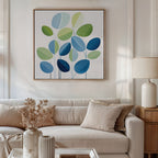 A modern living room features a light beige sofa, neutral pillows, a side table with a lamp, dried flowers in a vase, and the Layered Stillness Canvas Art—an abstract blue and green leaf painting—for stylish decor flair.
