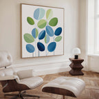 A modern living room with a white lounge chair and ottoman, wooden side table and lamp, and the Layered Stillness Canvas Art featuring blue and green leaf shapes. Light herringbone wood floors and a tall window complete the look.