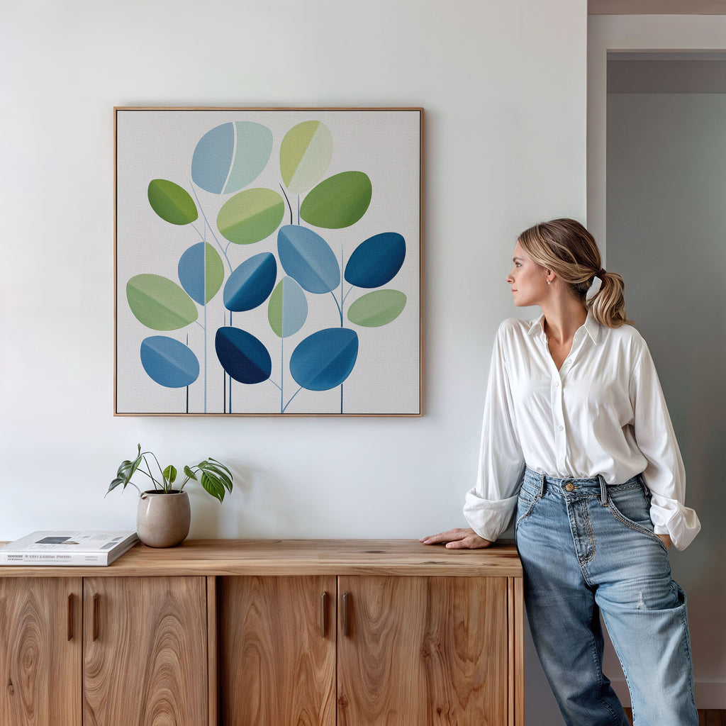 A woman in a white blouse and jeans leans against a wooden cabinet, admiring the Layered Stillness Canvas Art with green and blue leaves. A small potted plant and books on the cabinet complete the modern living room decor.