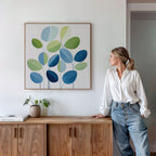 A woman in a white blouse and jeans leans against a wooden cabinet, admiring the Layered Stillness Canvas Art with green and blue leaves. A small potted plant and books on the cabinet complete the modern living room decor.