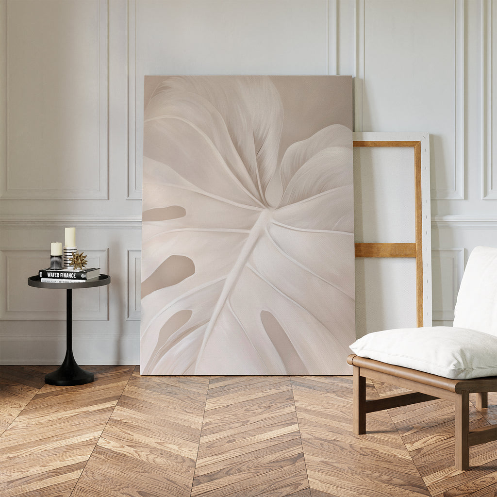 A minimalist room features the Leaf Whisper Abstract Canvas Art—a large pale leaf close-up—leaning on a white paneled wall beside an empty frame, with a black table holding candles and a wooden chair with a white cushion.