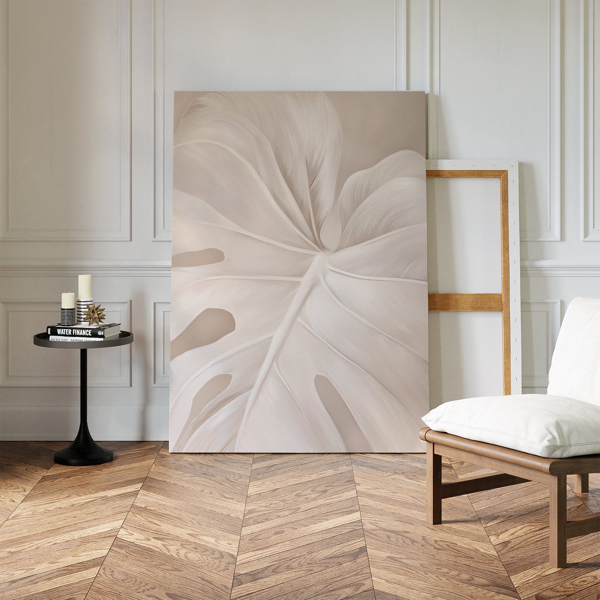 A minimalist room features the Leaf Whisper Abstract Canvas Art—a large pale leaf close-up—leaning on a white paneled wall beside an empty frame, with a black table holding candles and a wooden chair with a white cushion.