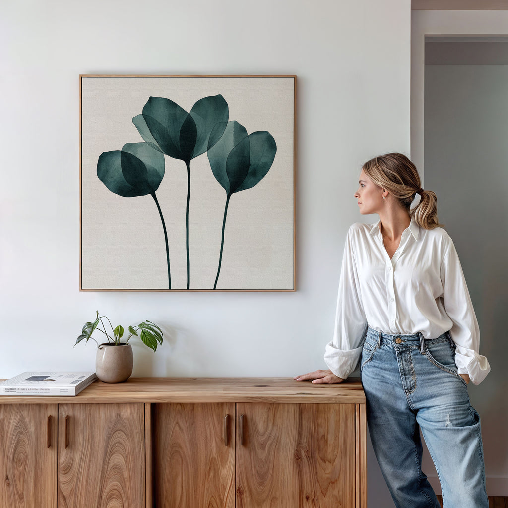 A woman in a white blouse and jeans leans against a wooden cabinet, admiring the Leafy Balance Canvas Art with green leaf designs above. A small potted plant and books complete the stylish living room decor.
