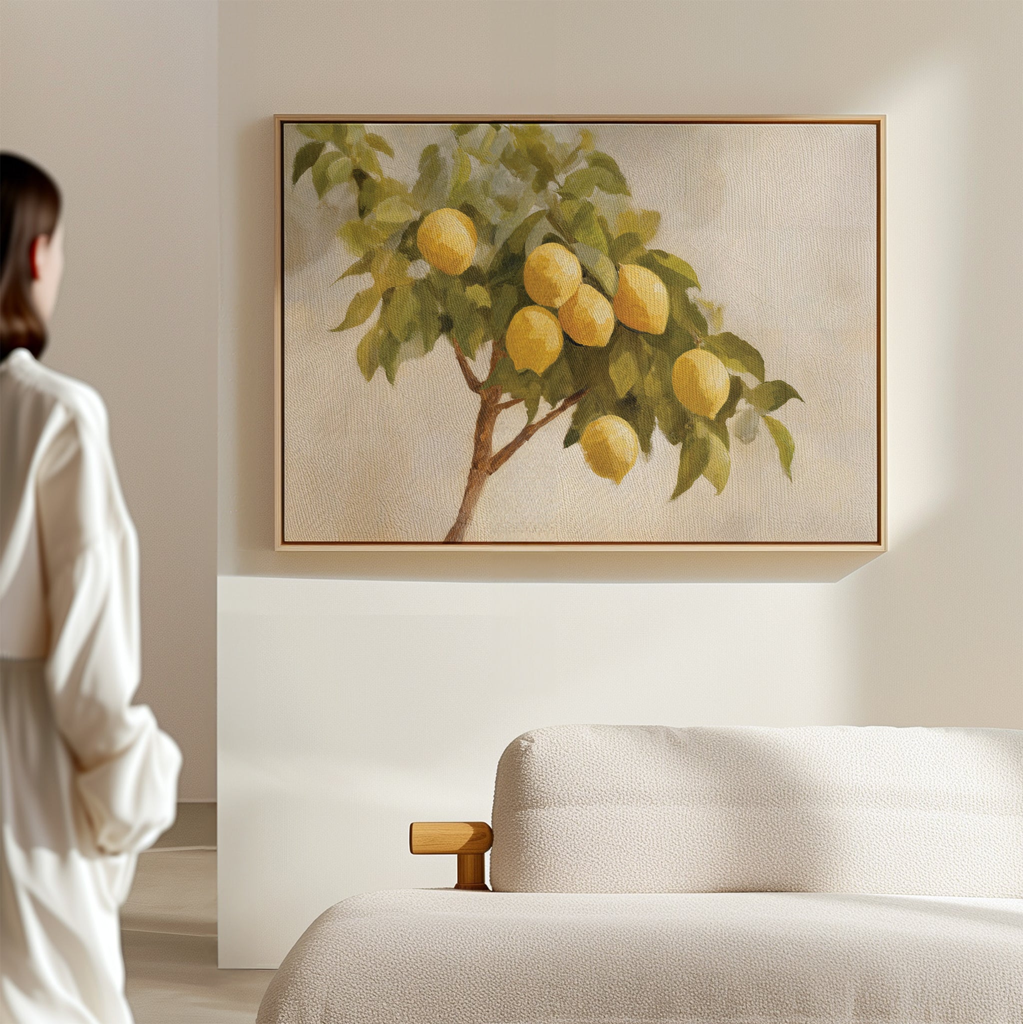A woman in a white robe stands in a minimalist room, gazing at Lemon Essence Canvas Art—a framed painting of a lemon tree with yellow lemons and green leaves—hanging above a light-colored sofa, ideal for Mediterranean kitchen decor.