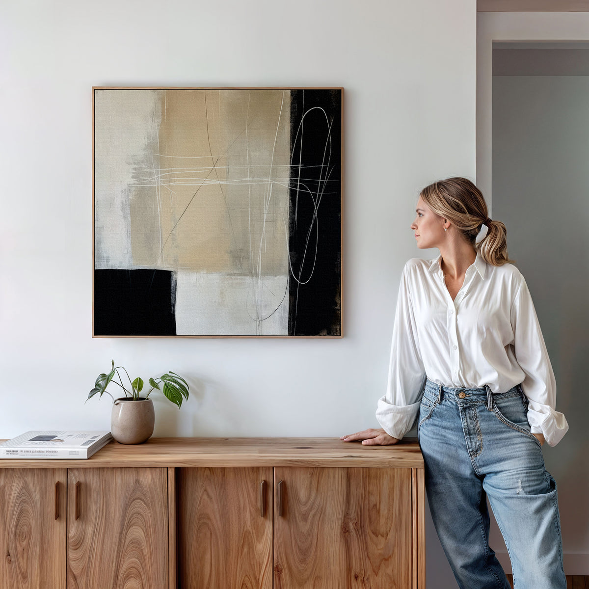 A woman in a white blouse and jeans leans against a wooden cabinet, admiring the Light and Shadow Abstract Canvas Art on the wall. A small potted plant and books rest on the cabinet in this modern, minimalistic living room.