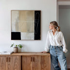 A woman in a white blouse and jeans leans against a wooden cabinet, admiring the Light and Shadow Abstract Canvas Art on the wall. A small potted plant and books rest on the cabinet in this modern, minimalistic living room.