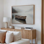 A cozy living room features a wooden sideboard with books, a lamp, and a bowl. Above, the peaceful Lonely Boat Canvas Art shows a rowboat on a misty lake. A light sofa is partially visible in the foreground.