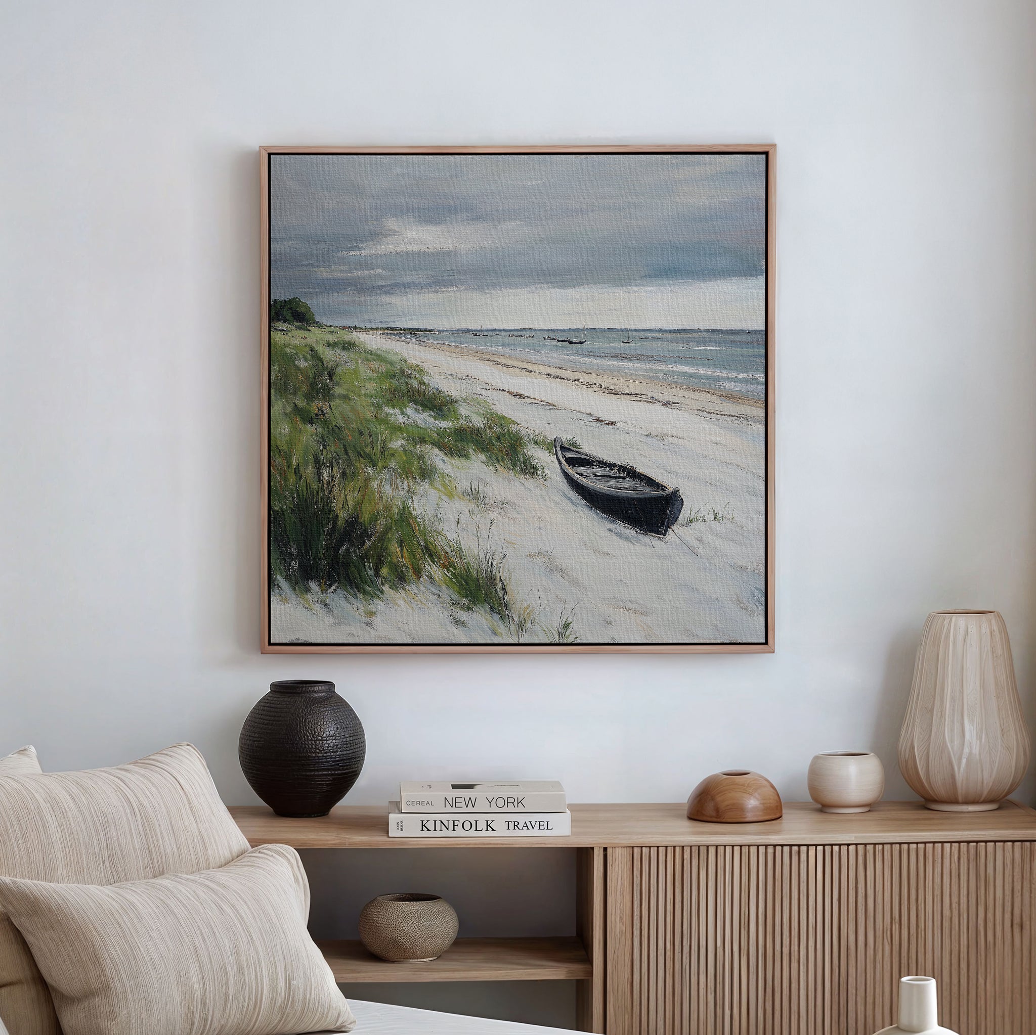 A living room features a beige sofa, neutral cushions, and a wooden sideboard with pottery and books. Above, the Lonely Coast Canvas Art, depicting a boat on a sandy beach with grasses and a cloudy sky, adds tranquil decor.