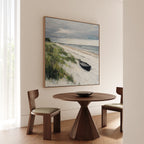 A round wooden table with two chairs sits in a bright room. Above, the Lonely Coast Canvas Art depicting a boat on sandy dunes adds tranquility as daylight filters through sheer curtains.