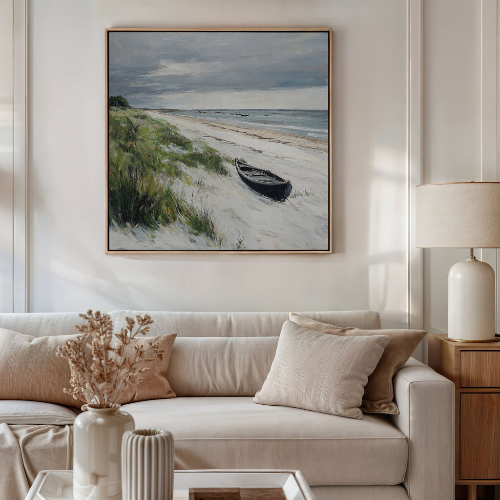 A cozy living room with a beige sofa, neutral pillows, a side table with a lamp, and dried flowers. Above the sofa hangs the Lonely Coast Canvas Art featuring a beach scene with grass, sand, a small boat, and cloudy sky.