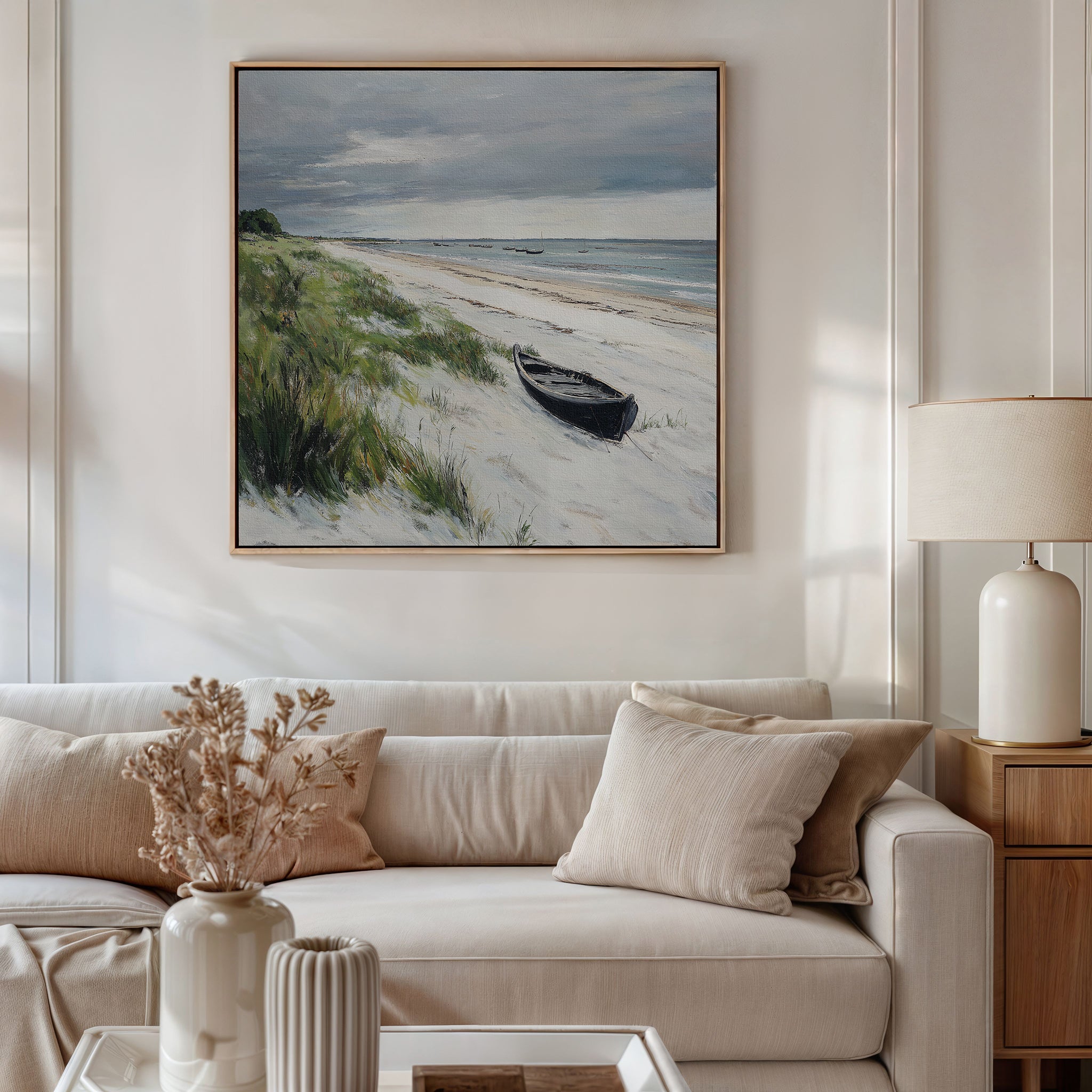 A cozy living room with a beige sofa, neutral pillows, a side table with a lamp, and dried flowers. Above the sofa hangs the Lonely Coast Canvas Art featuring a beach scene with grass, sand, a small boat, and cloudy sky.