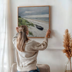 A woman with long blonde hair, in a beige sweater and jeans, hangs the Lonely Coast Canvas Art—a beach scene with a black boat—on a white wall in a cozy room with tranquil decor and pampas grass in a vase.