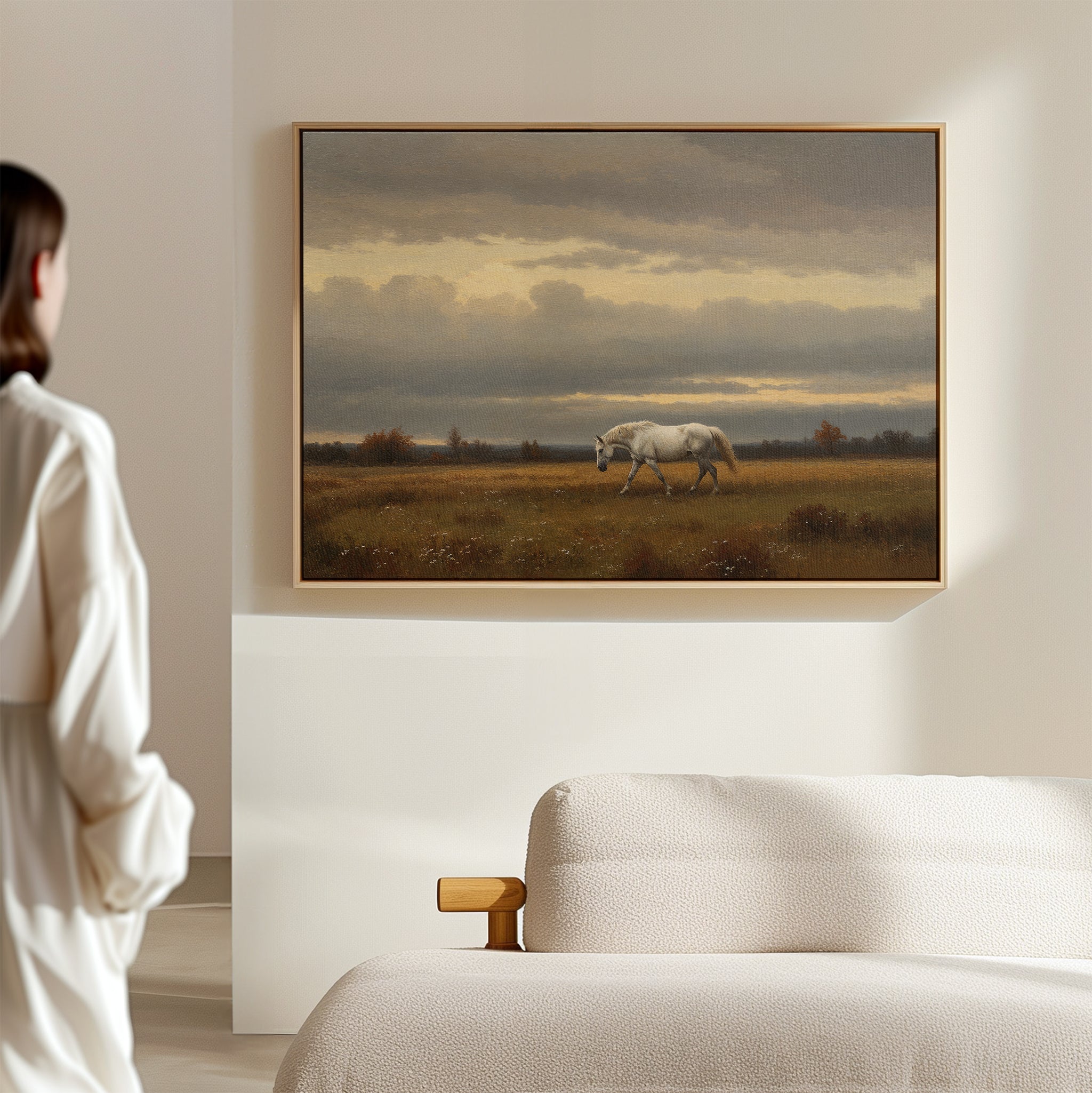 A person in white stands in a minimal living room, admiring the Low Light Walk Canvas Art—a countryside wall print featuring a white horse in a grassy field under a cloudy sky—hung above a beige sofa on a light wall.