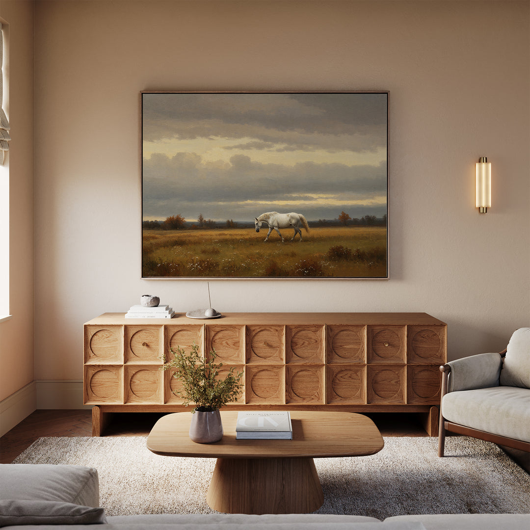 A cozy living room with neutral tones features a wooden sideboard, coffee table with book and vase, light sofa, and striking countryside wall art—Low Light Walk Canvas Art depicting a white horse in a field under a cloudy sky.
