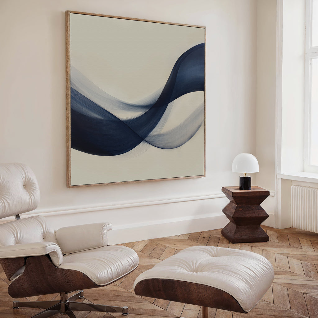 A modern living room with a white leather lounge chair and ottoman, wooden side table with a white lamp, and the minimalist Tidal Flow Canvas Art featuring abstract blue waves. Light from a tall window brightens the space.