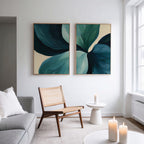 A modern living room with a gray sofa, wooden chair, round side table with a lit candle, and the "Lush Teal Foliage Set of 2" abstract canvas wall art. Sunlight from a large window enhances the contemporary decor.
