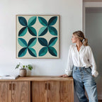 A woman in a white shirt and jeans leans against a wooden cabinet, gazing at the Marine Mosaic Canvas Art, which features an abstract petal design in teal and turquoise. A small plant and a book sit on the cabinet beside her.