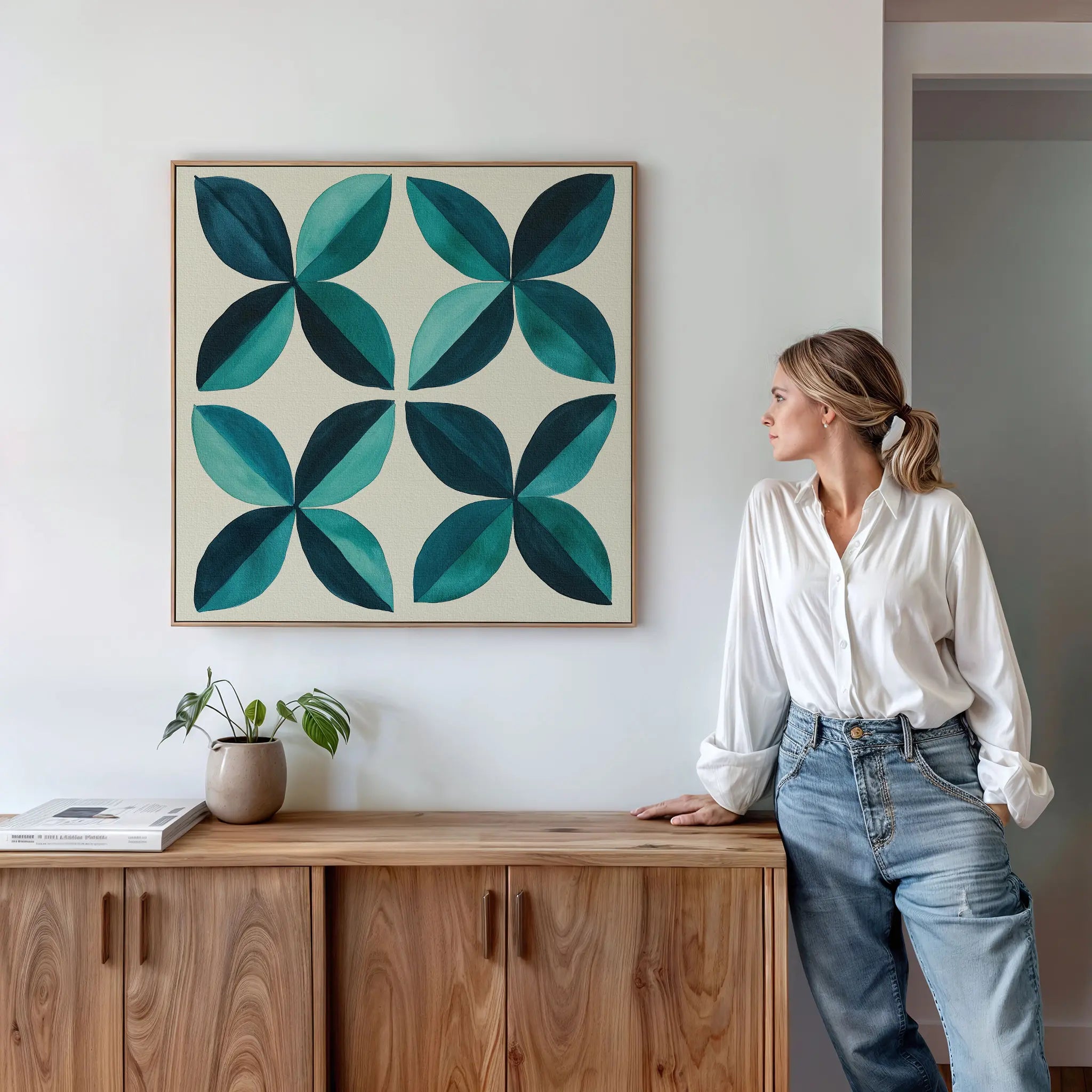 A woman in a white shirt and jeans leans against a wooden cabinet, gazing at the Marine Mosaic Canvas Art, which features an abstract petal design in teal and turquoise. A small plant and a book sit on the cabinet beside her.