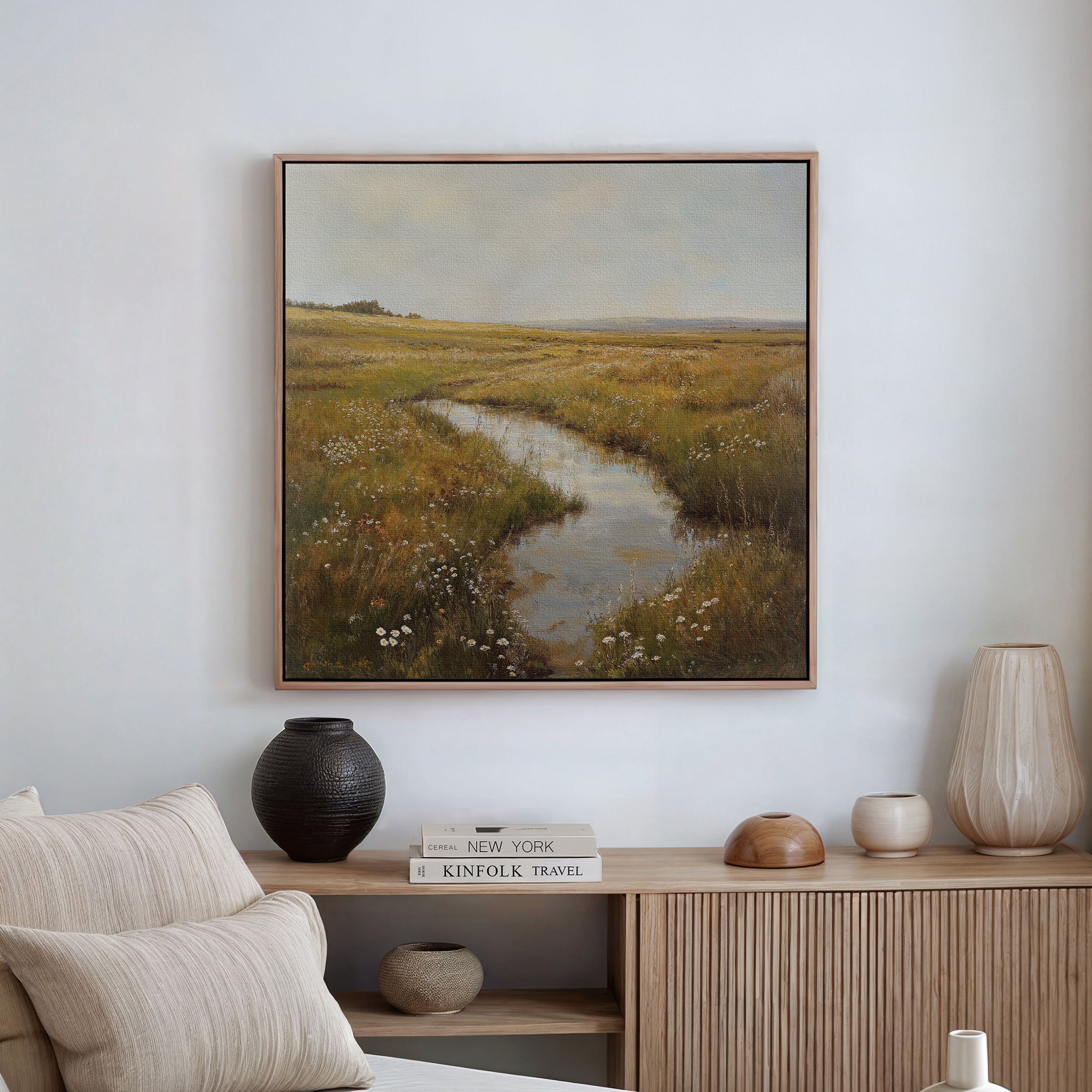 The Marshland Stream Canvas Art is framed and displayed above a light wood console with neutral decor—featuring a black vase, beige lamp, books, and a tan pillow on the sofa—in this minimalist living room.