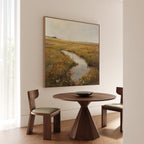 A round wooden table with two chairs sits in a bright room. Above it, the Marshland Stream Canvas Art depicting a grassy field and creek enhances the decor as sunlight filters through a sheer curtain.