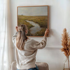 A woman with long blonde hair hangs the Marshland Stream Canvas Art—featuring a grassy field and river—on a white wall. Sunlight filters through curtains, while pampas grass in a vase adds to the stylish living room decor.