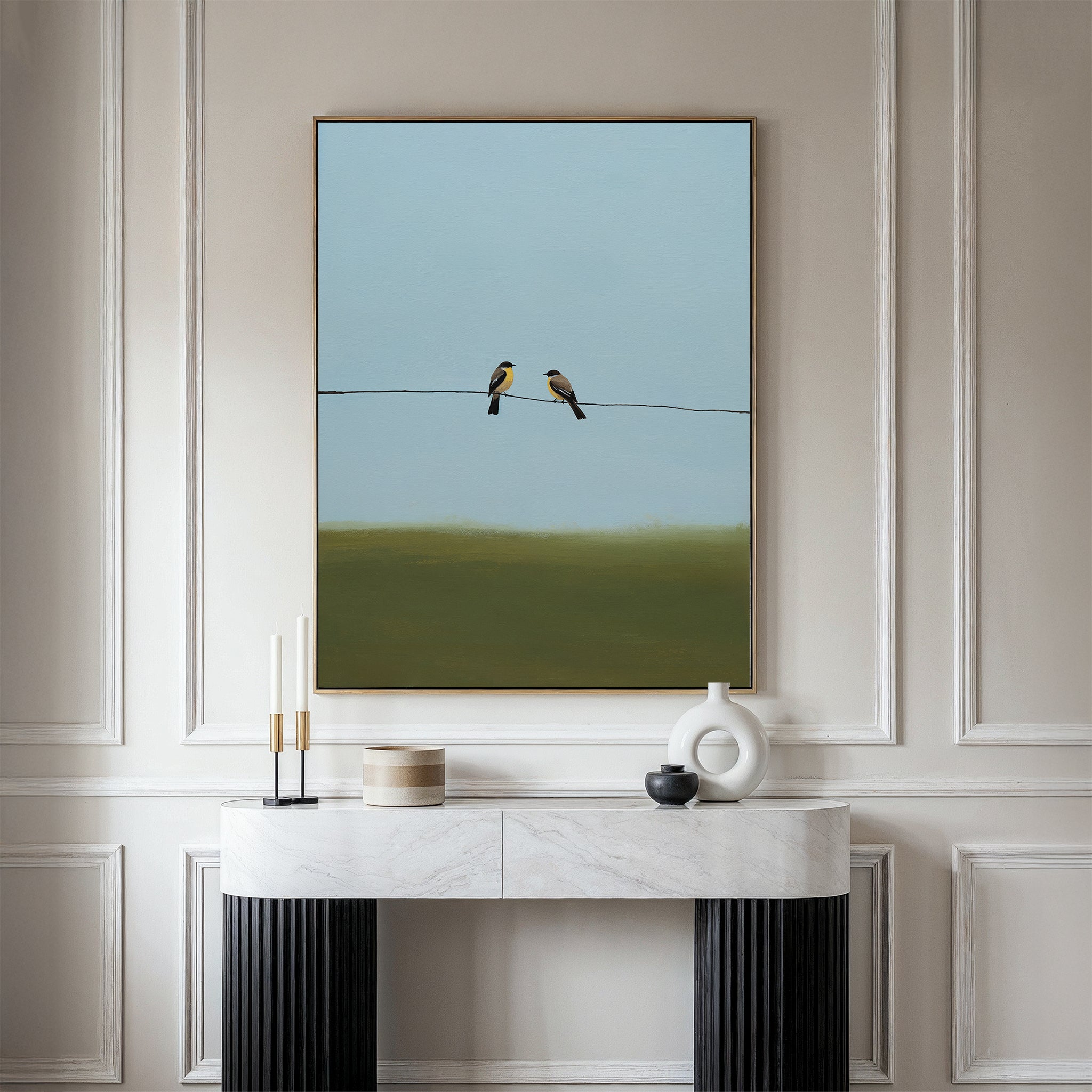 The Meadow Line Canvas Art, featuring two birds on a wire in a minimalist style, is displayed in a frame above a marble console table with candles and a white vase—ideal for enhancing modern, light-colored spaces.