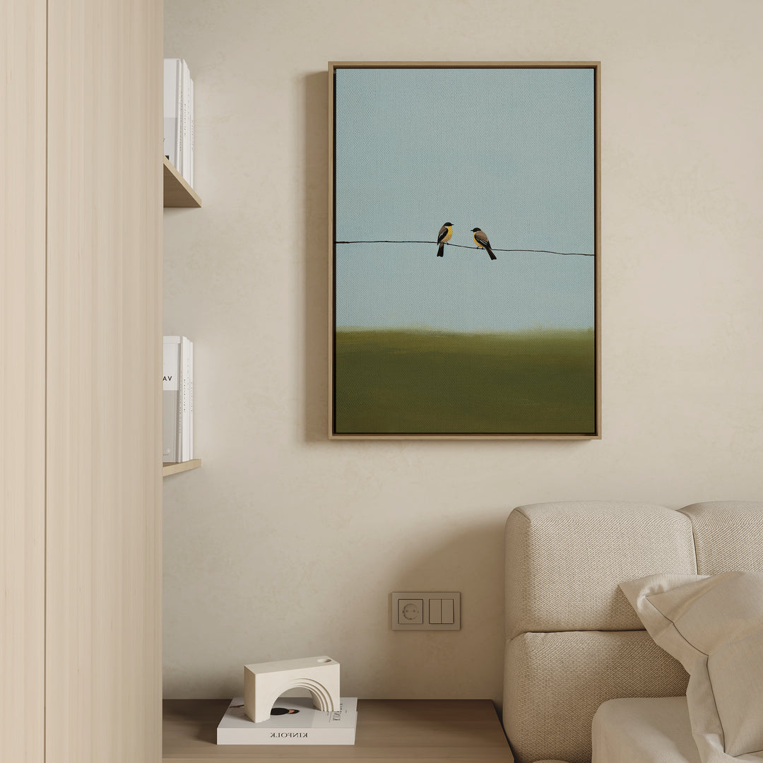 A minimalist living room features a beige couch, light wood shelves with books, and the Meadow Line Canvas Art—a framed print of two birds on a wire with a blue-green background—hanging on the wall.