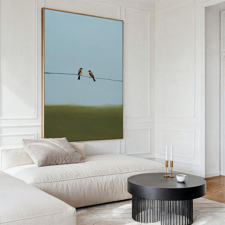 A modern living room highlights minimalist decor with the Meadow Line Canvas Art (two birds on a wire), a cream sofa with beige pillows, a round black coffee table with candles and cup, and white paneled walls.